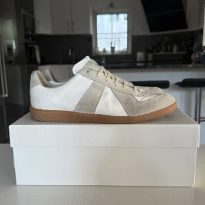 Maison Margiela sneakers i vitt och beige - Maison Margiela sneakers med klassisk siluett, vit och beige färgkombination, ovandel i skinn och mocka samt gummisula i brunt. Snygga detaljer med snörning och rund tå. Perfekta för dig som gillar stilrena och trendiga sneakers. Dom är använda en del men jag är säker på att det mesta går bort med lite vatten. Skriv om ni har frågor