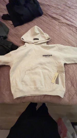 Essentials Fear of God vit hoodie oanvänd ny med prislapp - Säljer en snygg vit hoodie från Essentials Fear of God med svart logga på bröstet och ryggen. Tröjan har huva, känguruficka och ribbade muddar. Perfekt för dig som gillar streetwear och vill ha en clean look.