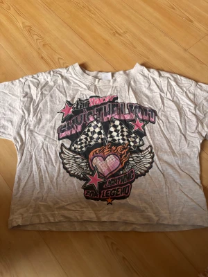 Vit t-shirt med tryck från Lindex - Cool vit t-shirt från Lindex med färgstarkt tryck på framsidan. Motivet har hjärta med eld, vingar, stjärnor och rutiga racingflaggor i rosa, svart och orange. Modellen är kortärmad och har en klassisk rund hals. Perfekt för dig som gillar statement-plagg.