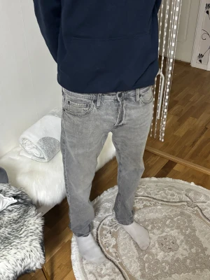 Grå tapered jeans från Jack & Jones - Säljer ett par grå jeans från Jack & Jones med snygg tvättad look och klassiska fem fickor. Modellen är tapered vilket ger en smalare passform nedtill. Jeansen har normal midja och är tillverkade i jeansmaterial med svarta detaljer på bakfickan.