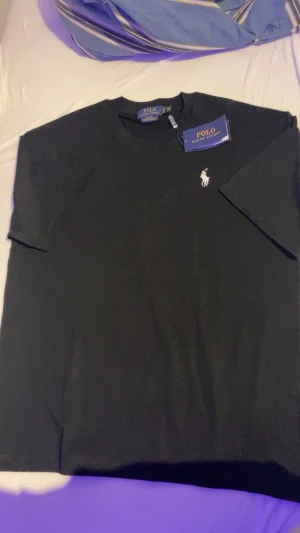 Svart t-shirt Polo Ralph Lauren - Svart t-shirt från Polo Ralph Lauren med klassisk vit broderad logga på bröstet. Slim fit-modell i mjuk bomull med rund halsringning. Enkelt och stilrent plagg som passar till allt.