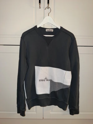 Mörkgrå sweatshirt från Stone Island - Cool mörkgrå sweatshirt från Stone Island med vit grafisk detalj och logga framtill. Tröjan har rund hals, långa ärmar och är gjord i mjuk bomull. Perfekt för dig som gillar streetwear och vill ha något som sticker ut.