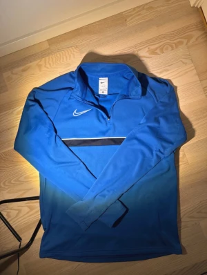 Blå Nike half zip tröja M - Snygg blå långärmad tröja från Nike med half zip och Dri-Fit material. Tröjan har en vit Nike-logga på bröstet och en svart/vit rand över magen. Perfekt för träning eller chill, med sportig vibe och skön passform.
