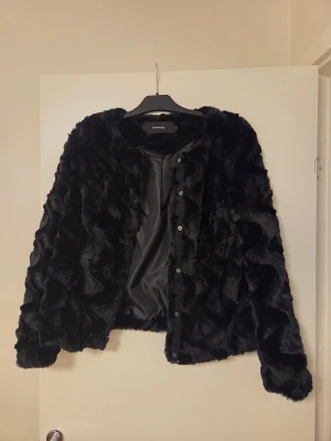 Svart pälsjacka från Vero Moda - Säljer en svart pälsjacka från Vero Moda med snyggt vågigt mönster i pälsen. Jackan har rund hals, långa ärmar och knäppning med knappar framtill. Perfekt för dig som vill ha en mjuk och fluffig look med lite edge.