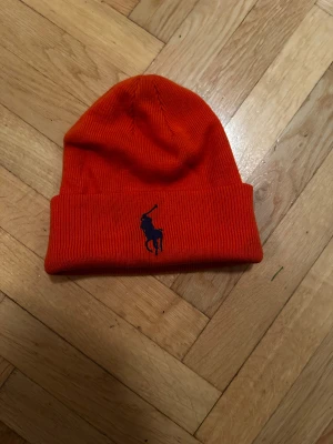 Orange mössa från Polo Ralph Lauren - Snygg orange mössa från Polo Ralph Lauren med klassisk blå broderad logga framtill. Perfekt för kalla dagar. Pris gör att diskutera
