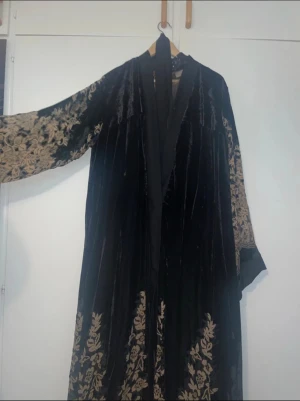 Svart långklänning med blommigt mönster - Unik svart långklänning i sammet med beige blommigt mönster längs ärmar och nederkant. Klänningen har lång ärm och en elegant omlottkrage. Perfekt för dig som gillar bohemisk och lyxig stil med detaljer som sticker ut. Aldrig använt, var en present till morsan men hon tyckte inte om färgen svart. Köpte den från Irak men den är gjord i Förenade arabemiraten. Köpte den för 1500kr men säljer den för 799kr Storlek 66 men den är oversize så att den passar varje storlek till exempel XL, L, M