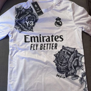 Vit Real Madrid Y-3 x Adidas fotbollströja - Säljer en vit Real Madrid fotbollströja från Adidas Y-3 och Yohji Yamamoto. Tröjan har korta ärmar, svarta ros-motiv, Real Madrid-logga och Emirates Fly Better-tryck på bröstet. Materialet är lätt och andas, perfekt för match eller träning.