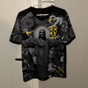 Nike Brasil svart grafisk fotbollströja - Unik svart Brasilien fotbollströja från Nike med detaljerat grafiskt tryck av Kristusstatyn och brasilianska symboler. Gula och gråa accenter, CBF-emblem på bröstet och ryggen. Tillverkad i lätt, andningsbart material. Perfekt för dig som vill sticka ut på planen eller läktaren.