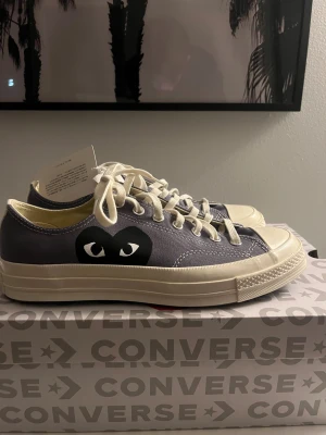 Converse x Comme des Garçons Play sneakers - Säljer ett par sprillans nya Converse x Comme des Garçons Play sneakers i mörkgrå canvas med vit tå och sula. Skorna har klassisk snörning och det ikoniska svarta hjärta med ögon på sidan. Perfekt för dig som gillar coola samarbeten och streetstyle.