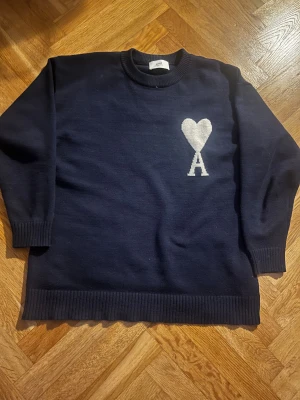 Mörkblå stickad tröja från Ami - Snygg mörkblå stickad tröja från Ami med vit hjärtlogga och bokstaven A på bröstet. Tröjan har rund hals, ribbade muddar och lång ärm. Perfekt för dig som gillar stilrena detaljer och vill ha något som sticker ut lite extra.