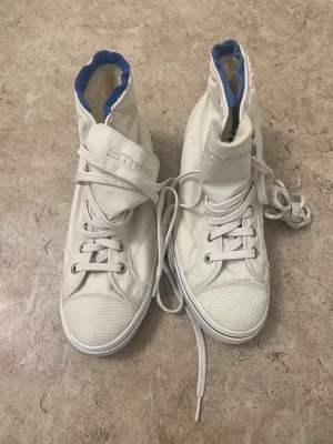 Vita höga sneakers från Tretorn - Säljer ett par vita höga sneakers från Tretorn med snörning och rund tå. Skorna har en mjuk och varm ullfoder på insidan med blå detaljer runt kanten. Yttermaterialet är slitstark textil och sulan är platt. Perfekta för kyliga dagar.