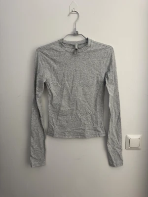 Grå långärmad SLIM FIT topp från SKIMS storlek XS  - Säljer en enkel och stilren grå långärmad topp från SKIMS i storlek XS. Toppen har rund hals och är gjord i en mjuk blandning av bomull, polyester och elastan för en skön passform. Perfekt att styla med jeans eller kjol för en clean look.