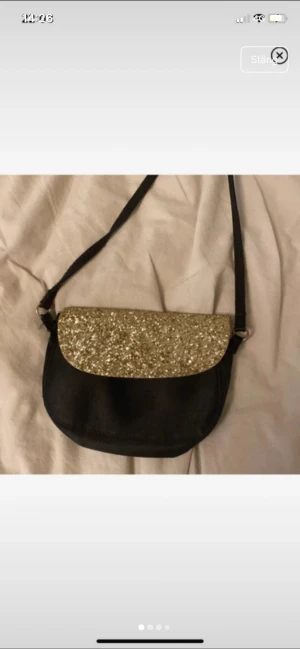 Svart axelväska med glittrigt lock - Svart axelväska med rundad form och justerbar rem. Locket är täckt av guldglittrigt material som ger en extra cool touch. Väskan stängs med magnetknapp och har plats för det viktigaste. Perfekt för dig som gillar att sticka ut med detaljer.