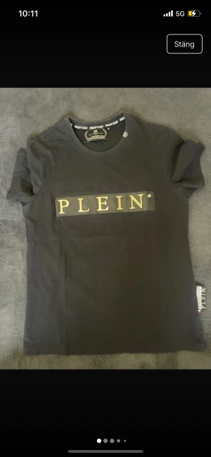 Svart Philipp Plein t-shirt med guldtryck - Snygg svart t-shirt från Philipp Plein med guldigt PLEIN-tryck på bröstet. Rund hals, korta ärmar och klassisk passform. Materialet är mjuk bomull och har en exklusiv känsla med märkets logga i nacken och på etiketten vid sidan.