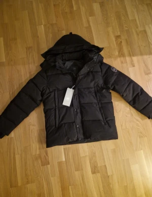 Svart jacka från Canada Goose - Svart jacka från Canada Goose med huva, quiltad design och logotyp på ärmen. Jackan har dragkedja och knappar framtill, samt två stora fickor.. Snygg och stilren modell som håller dig varm hela vintern. Helt ny!