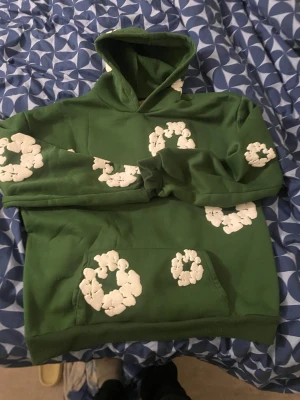 Grön hoodie från Denim Tears - Säljer en grön hoodie från Denim Tears. Hoodien har huva, magficka och ribbade muddar. Perfekt statement piece för dig som vill sticka ut med cool design.