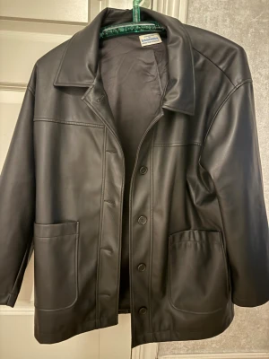 Svart skinnjacka från Barbara - Snygg svart skinnjacka (imitation) från Barbara med klassisk krage och stora framfickor. Jackan har knäppning med knappar framtill och en rak, något loose passform. Perfekt för dig som vill ha en stilren och tidlös look.