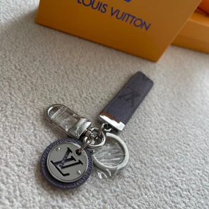  Vuitton nyckelring i silver och grått - Exklusiv  Vuitton nyckelring med silverfärgade metalldetaljer och grå läderrem. Rund bricka med -logga i silver och grått, samt präglad text. Snygg och stilren accessoar med klassisk design och lyxig känsla.