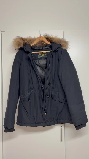 Svart/marinblå jacka från Woolrich med päls - Svart/marinblå jacka från Woolrich med huva med äkta päls. Jackan har både dragkedja och knappar framtill, två stora fickor med knappar och är tillverkad i en mix av bomull och nylon. Perfekt inför vintern med massa snö och minusgrader! Jackan är helt ny utan prislapp och ligger idag runt 8000kr. Jackan säljs då den inte kommer till användning, priset kan diskuteras!!!