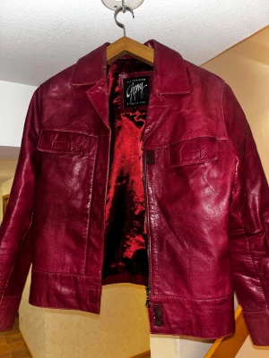 Vinröd skinnjacka från Gipsy  - Passar Medium - Säljer en vinröd skinnjacka från Gipsy i storlek XL men den passar M. Jackan har klassisk krage, två bröstfickor med lock och dragkedja framtill. Materialet är äkta skinn med glansig röd insida. Säljer för jackan är för liten. Liten defekt sen bild 3