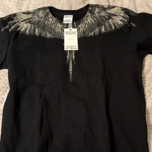 Svart tröja Marcelo Burlon med vingar - Cool svart tröja från Marcelo Burlon County of Milan med ikoniskt vingtryck i grått över axlar och hals, både fram och bak. Tröjan har rund hals och lång ärm, tillverkad i mjuk bomull. Perfekt för dig som gillar streetwear med unik design.