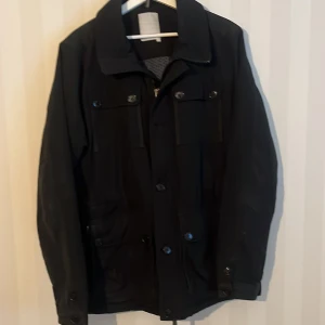 Svart fieldjacka från J.Lindeberg - Svart vindjacka från J.Lindeberg med klassisk krage, knappar framtill och flera fickor. Jackan har ett snyggt mönstrat innerfoder i blått och svart samt diskret logopatch på ärmen. Perfekt för dig som vill ha en stilren och funktionell jacka.