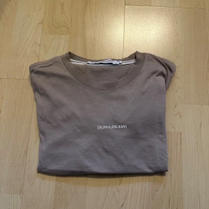 Calvin Klein beige t-shirt - T-shirt från Calvin Klein Jeans med en rund halsringning och korta ärmar. Den har en enkel design med en liten logotyp tryckt på bröstet.