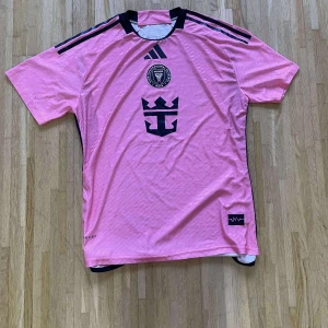 Inter Miami CF rosa matchtröja Adidas - Säljer en rosa Inter Miami CF fotbollströja från Adidas med svarta detaljer och klubbmärke på bröstet. Tröjan har korta ärmar, svart krage och svarta ränder på axlarna. Tillverkad i lätt funktionsmaterial, perfekt för träning eller match.