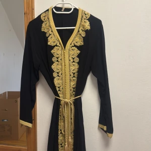 Svart långklänning med guld brodyr - Svart långklänning med lyxig guld brodyr längs hela framsidan, ärmslut och nederkant. Klänningen har v-ringning, långa ärmar och ett matchande guldigt knytbälte i midjan. Snygga slitsar nertill och detaljer med små glittrande stenar för extra bling.