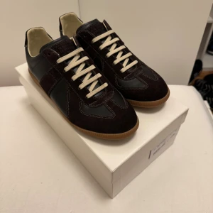 Maison Margiela Gats  - Maison Margiela Gats i mörkbrunt med kontrasterande vita snören och beige insida. Ovandel i lammskinn och kalvskinn, detaljerad med mocka och läderpaneler. Klassisk gummisula med Maison Margiela-logga undertill. Snygg retrostil och rund tå. Skorna är helt nya och oanvända pris 1300kr vilket är jätte billigt prisen kan diskuteras vid snabb affär. 