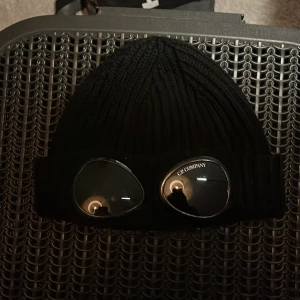 Svart goggle-mössa från C.P. Company - Svart ribbstickad mössa från C.P. Company med ikoniska goggle-linser framtill. Mössan är tillverkad i ull och har en uppvikt kant. Perfekt accessoar för dig som vill sticka ut med streetwear-vibe.