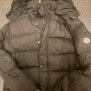 Moncler vezere  - Säljer en svart dunjacka från Moncler med klassisk pufferdesign. Jackan har huva, dragkedja och knappar framtill samt Moncler-logga på ärmen. Perfekt för kalla dagar och riktigt snygg till streetwear-stilen.