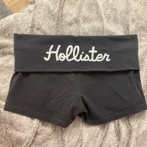 Svarta Hollister shorts med logga - Svarta shorts från Hollister med bred, vikt midja och stor vit broderad Hollister-logga framtill. På baksidan finns en liten röd logga och siffran 1922. Mjukt och stretchigt bomullsmaterial, perfekt för chill eller träning.