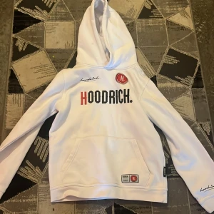 Vit hoodie från Hoodrich med tryck - Säljer en vit hoodie från Hoodrich med stor logga framtill och coolt tryck på ryggen. Tröjan har luva, magficka och detaljer i svart och rött. Perfekt för dig som gillar streetwear och vill sticka ut med snygga prints.