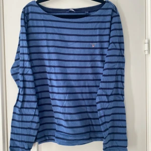 Blå randig långärmad tröja GANT XL - Snygg blå långärmad tröja från GANT i storlek XL. Tröjan har marinblå och ljusblå ränder, rund hals och ett litet broderat GANT-logo på bröstet. Tillverkad i mjuk bomull som är skön mot huden. Perfekt för dig som gillar klassisk och chill stil. Mått Över bröstet: ca 58 cm Längd från axeln och ner: ca 60 cm  100% bomull  Kommer från ett djur och rökfritt hem.