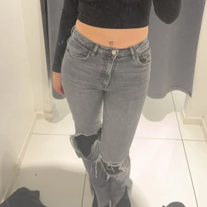 Grå ripped jeans med bootcut - Säljer ett par grå jeans med bootcut och stora slitningar vid knäna. Jeansen har klassisk femficksdesign, knapp och dragkedja framtill samt en snygg, lite tvättad look. Perfekta för dig som gillar en edgy stil.