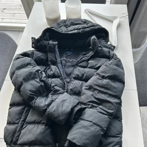 Svart pufferjacka från GANT - Svart pufferjacka från GANT med huva och dragkedja framtill. Jackan har ett stilrent, quiltat mönster och logotypen broderad på bröstet. Perfekt för kalla dagar. Den är väldig skön och har en perfekt passform. Den har inte några hål eller skador, som ny. Passar dig som är mellan 170-182cm lång. Priset går att diskutera.