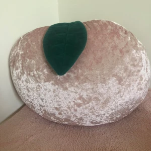 Persikoformad kudde - Bilderna visar en plyschkudde i form av en persika med ett grönt blad, tillverkad i mjukt, glansigt tyg. 