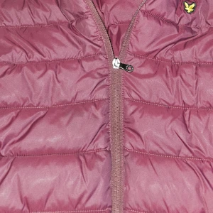 Vinröd dunväst från Lyle & Scott Junior - Säljer en vinröd dunväst från Lyle & Scott Junior med gul logga på bröstet. Västen har quiltad design, dragkedja framtill och är tillverkad i syntetmaterial. Perfekt för lager-på-lager under höst och vinter. Passar dig som gillar stilren och sportig look.