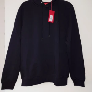 Svart hoodie från HUGO med snörning - Svart hoodie från HUGO med stilrena detaljer och snörning i huvan. Tröjan har lång ärm, ribbade muddar och diskret HUGO-logga på snörändarna. Mjuk bomullskvalitet som känns skön mot huden. Perfekt för en chill och snygg look.