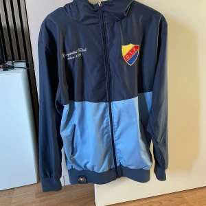 Djurgården vindjacka blå Stl. S - Djurgården IF vindjacka i två nyanser av blått med klubbmärke på bröstet och broderad text. Jackan har huva, hel dragkedja och två fickor med med dragkedja. 