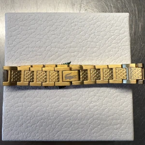 Lacoste länkarmband i guld  - Stilrent länkarmband från Lacoste med ikoniska krokodil-loggan. Finns i guldfärgärg, med snyggt mönstrade länkar som ger en cool och modern känsla. Perfekt accessoar för att lyfta din outfit med en sportig touch.