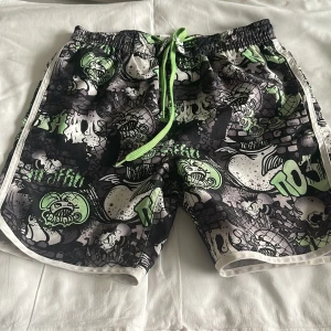 Badshorts med grön graffiti från KappAhl - Säljer coola badshorts från KappAhl med grymt graffitiinspirerat mönster i svart, grått, vitt och grönt. Shortsen har elastisk midja med grön snörning och vita detaljer längs sidorna. Perfekta för stranden eller poolen!