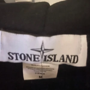 Svart hoodie från Stone Island M - Svart hoodie från Stone Island med dragkedja framtill, huva med snörning och klassisk logotyp-patch på vänster ärm. Tröjan har känguruficka och ribbade muddar. Perfekt för dig som gillar streetwear och vill ha en clean look. Passar M-S