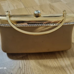 Guldig kuvertväska med strassdetaljer - Snygg kuvertväska i guld med glittrande strass längs kanten och ett elegant handtag i metall. Väskan har en klassisk rektangulär form och ett dekorativt lås med rund sten på toppen. Perfekt för att lyfta din outfit med lite extra bling.