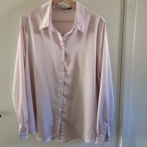 Ljusrosa skjorta från Boohoo - Snygg ljusrosa skjorta från Boohoo med klassisk krage och knäppning framtill. Skjortan har långa ärmar och är tillverkad i ett glansigt, mjukt material som ger en lyxig känsla. Perfekt för dig som gillar stilrena och enkla plagg.