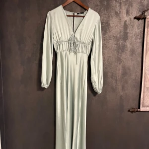 Ljusgrön långklänning med snörning - Snygg ljusgrön långklänning med v-ringning och snörning framtill. Klänningen har långa ärmar med lätt puff och är tillverkad i ett glansigt, mjukt material som ger en lyxig känsla. Perfekt för dig som gillar stilrena och eleganta plagg.