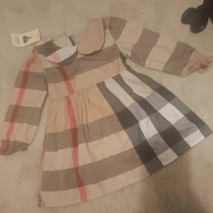 Rutig kortklänning  - Supersöt rutig klänning  i klassiska beige, bruna, svarta och röda toner. Klänningen har långärmade puffärmar, rund krage och ett mjukt bomullsmaterial. Perfekt för dig som gillar ikoniska mönster och stilren design.