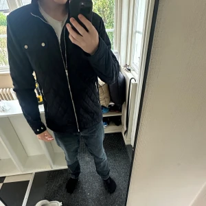 Svart quiltad jacka från Selected Homme - Snygg svart quiltad jacka från Selected Homme med dragkedja framtill, två bröstfickor med knappar och ståkrage. Jackan har en stilren look och är perfekt för dig som gillar en klassisk men modern stil.