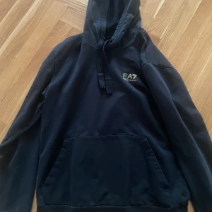 Svart hoodie från EA7 Emporio Armani - Svart hoodie från EA7 Emporio Armani med gul logga på bröstet och dekorativa gula ränder på ryggen. Klassisk känguruficka framtill, huva med snörning och långa ärmar. Perfekt för en stilren och sportig look.